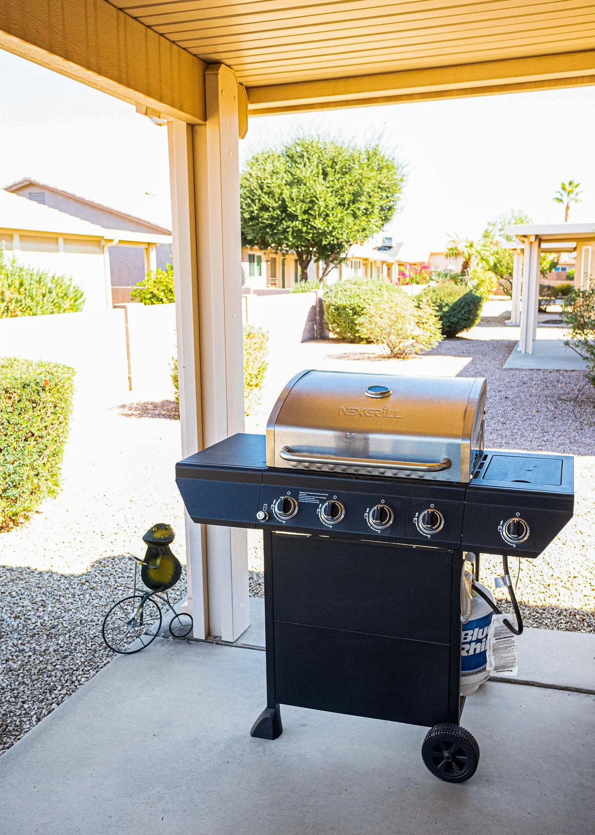 A relaxed backyard setting is depicted, with a prominent BBQ grill in the foreground, attractive landscaping, and a decorative outdoor lamp. The area is designed for barbecuing, outdoor dining, and socializing, emphasizing a sunny and cheerful mood.