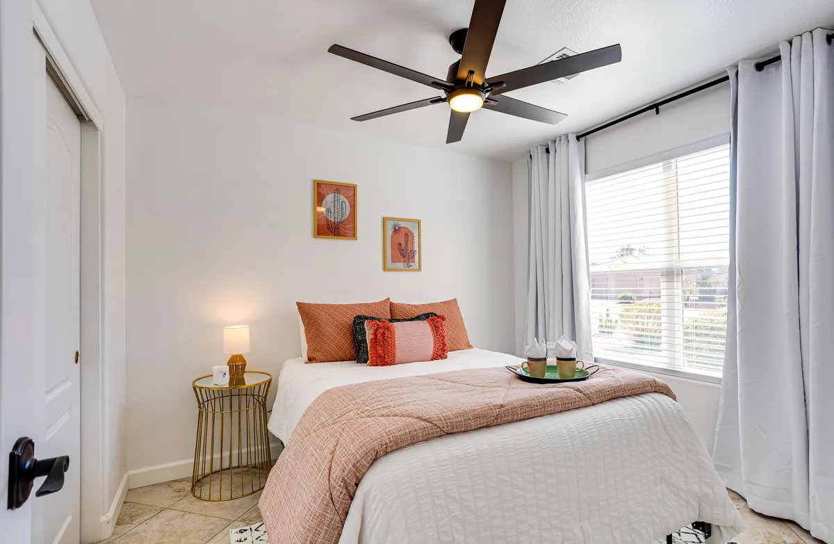 A bright, inviting bedroom with a plush bed as the centerpiece, complemented by decorative artwork and a side table with a lamp. Natural light pours in through the window, creating a calm and restful ambiance.