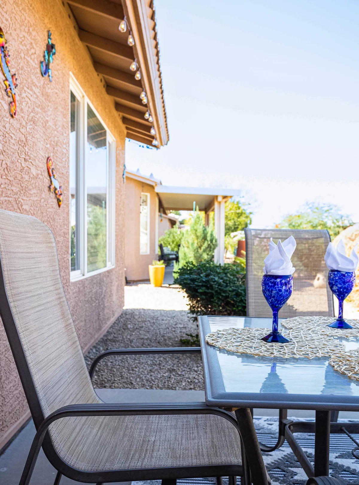 An inviting outdoor seating area with a well-set table, blue glasses, and decorative wall art, bathed in natural sunlight. The setup is perfect for group relaxation, informal meals, or club meetings, capturing a sense of vibrant social life.