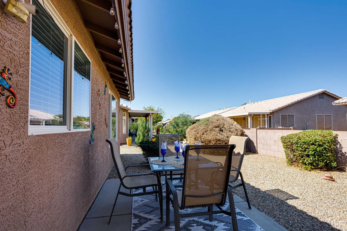 An inviting exterior patio setup featuring a dining table set with blue glasses, decorative wall art, and lush landscaped shrubs in the background. The space feels open, social, and ideal for outdoor gatherings or relaxation.