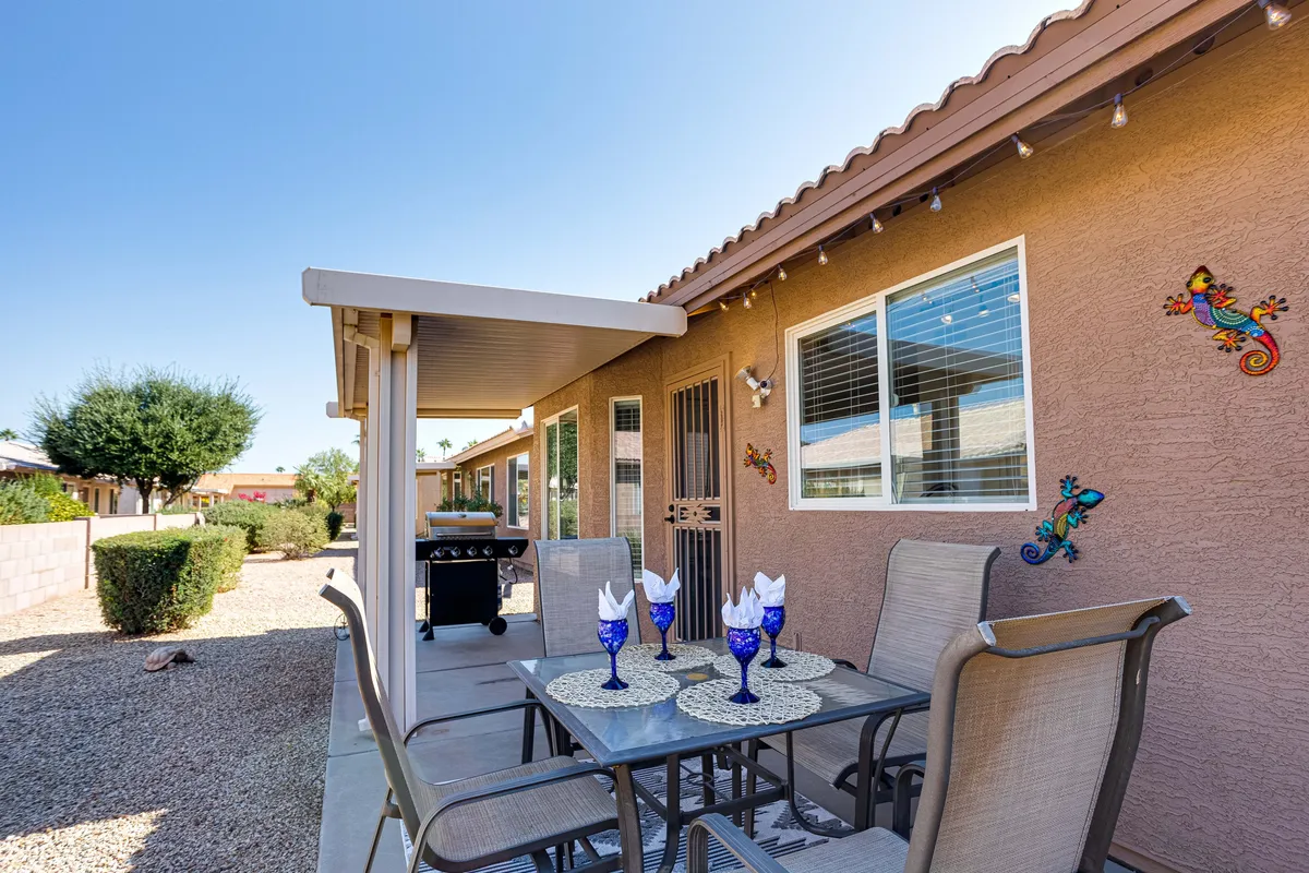 This image captures a vibrant backyard scene featuring a table set with blue glasses, decorative wall art, and beautifully landscaped gardens. The outdoor dining area is inviting, surrounded by lush greenery, and clearly designed for social gatherings, sunset dinners, or relaxed mornings.