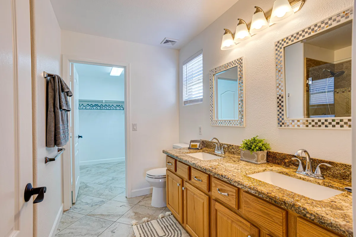 A spa-like bathroom with a double sink vanity, granite countertop, large mirrors, and a visible walk-in closet door. The space is bright, organized, and modern, exuding a sense of tranquility and convenience.