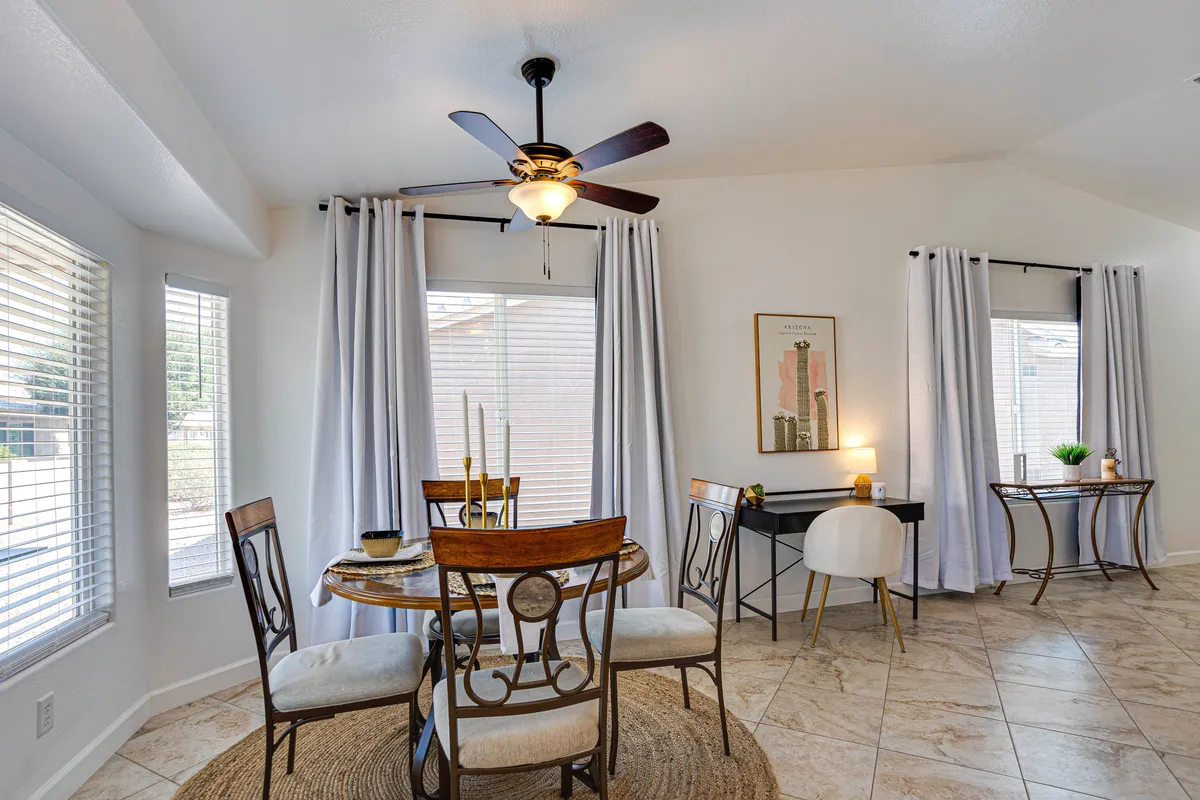 A bright, welcoming dining area with a dining table and chairs set near sunny windows with curtains, complemented by a desk and chair in the background and decorative artwork that adds character to the space. The ambiance suggests an inviting spot for meals, gatherings, or remote work in a comfortable home setting.