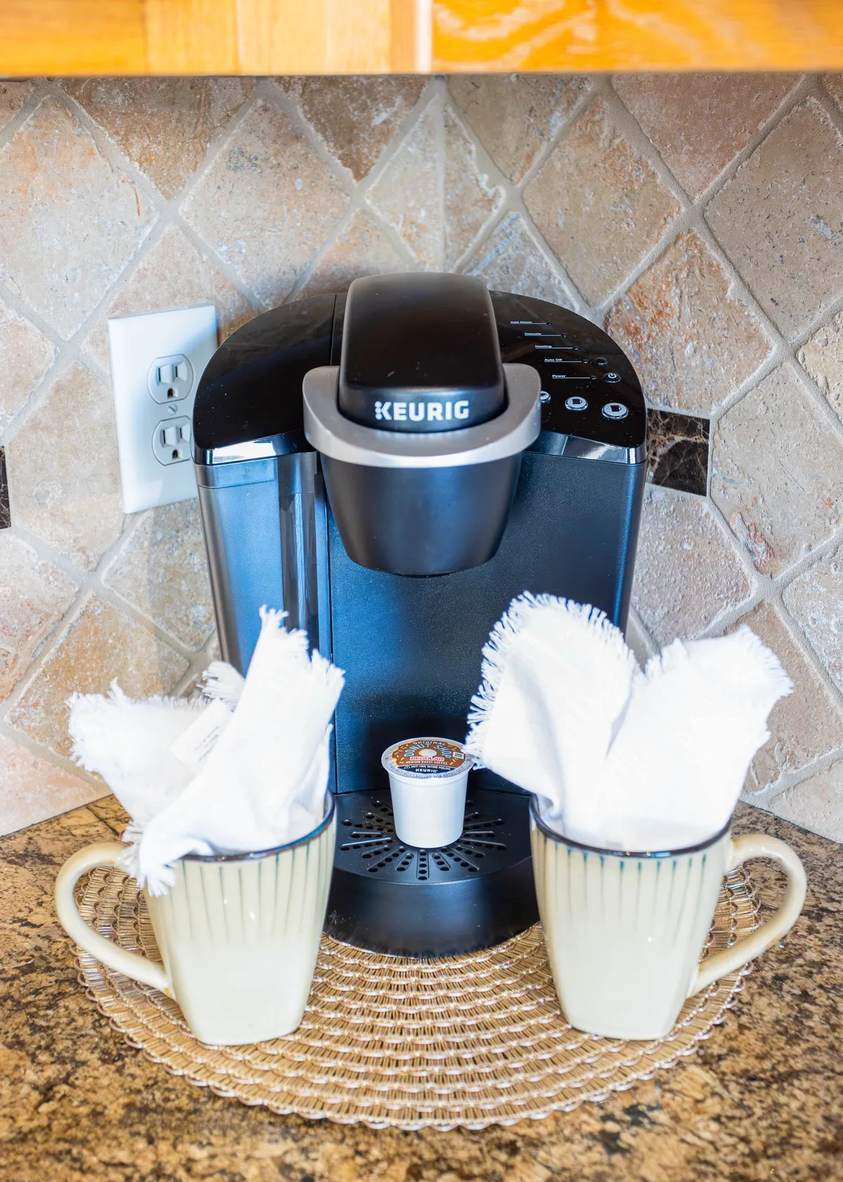 A homey coffee nook with a Keurig machine, two cups, a coffee pod, and a woven placemat, set on a kitchen counter. The scene radiates morning comfort and casual interaction, indicating places where residents can start their day or enjoy casual conversation.