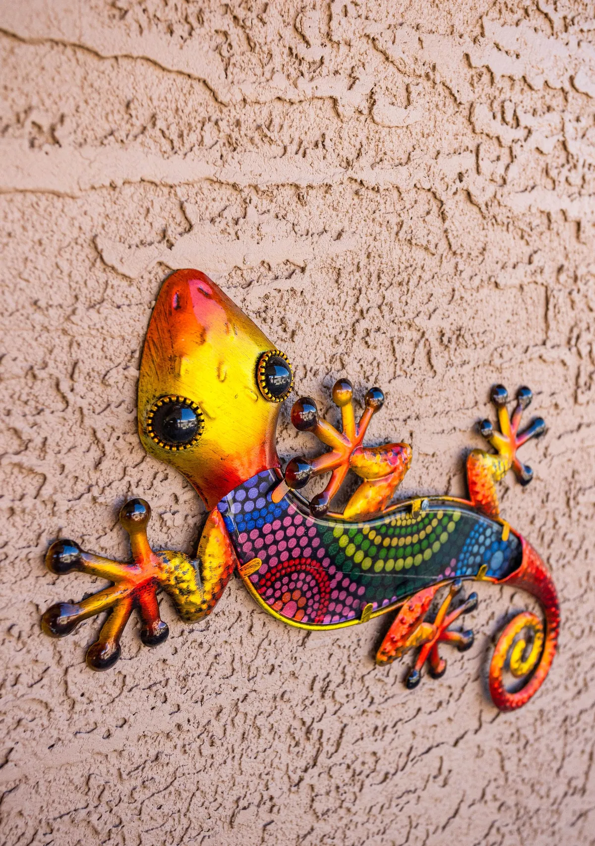 A close-up of a colorful metal gecko decoration mounted on a textured exterior wall. The bright and playful decor adds a cheerful, welcoming touch to the outdoor environment.