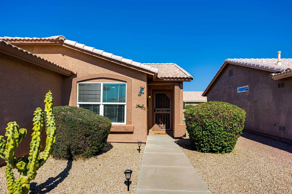 The image features a front door framed by desert landscaping, including a cactus, bushes, and decorative lizards, creating a distinctly Southwestern sense of place. There's an inviting sense of transition between indoors and outdoors, hinting at local vibes and the spirit of exploration.